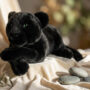 Peluche panthère noire allongée aux yeux verts sur tissu crème avec galets décoratifs, ambiance zen relaxante